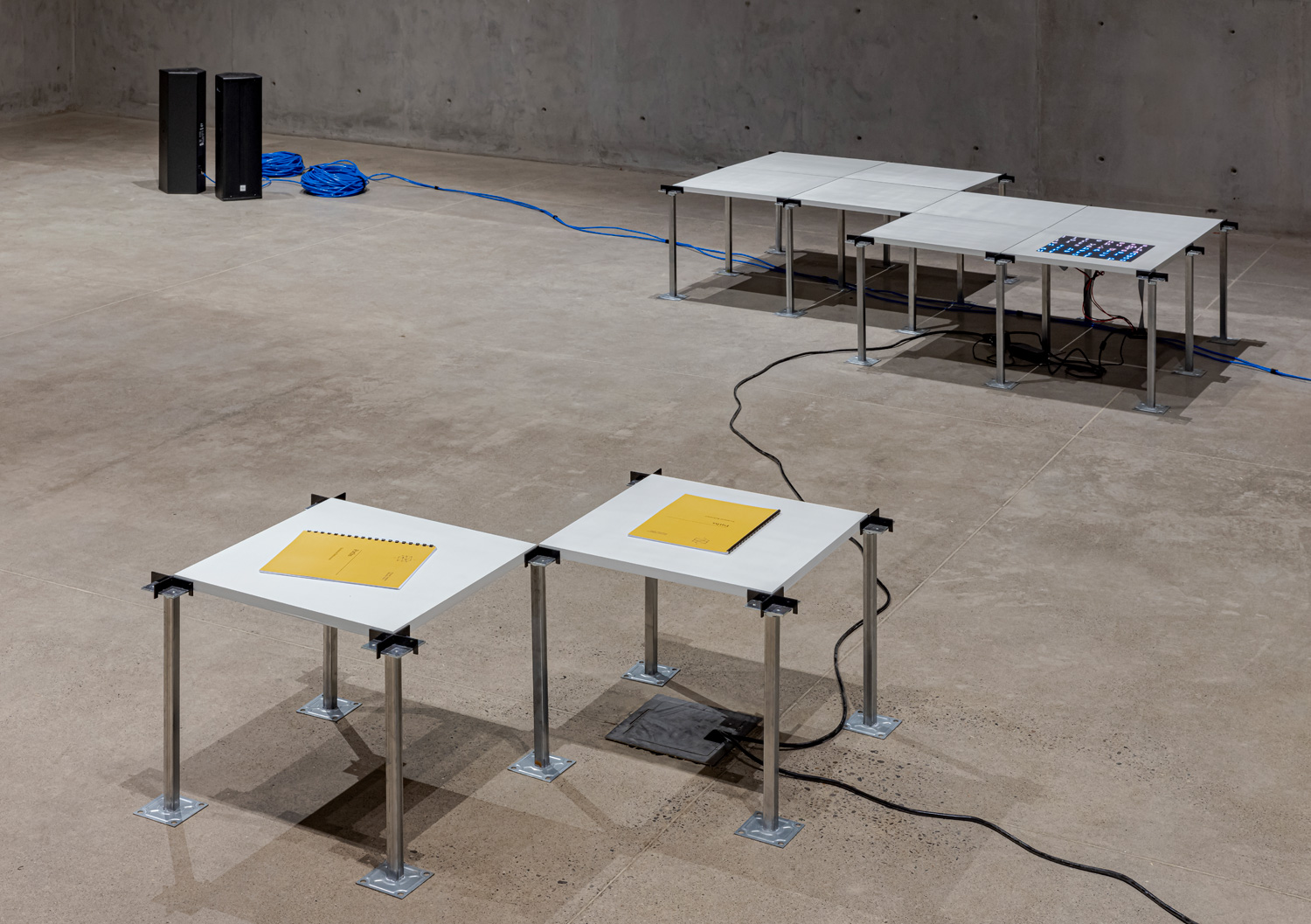 A closeup of one of the modular platforms. Sitting on it are two copies of 'Paths', the exhibition book. It has a yellow cover and black comb binding. In the background is another platform with an embedded LED matrix display. Next to that are two tall speakers sitting on the ground, with two large spools of blue ethernet cable.