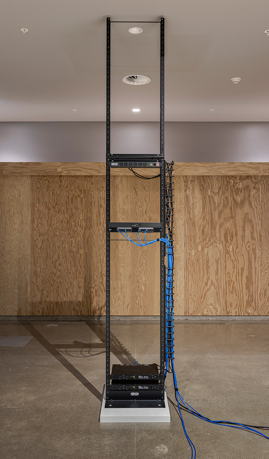 A modified server rack that extends to the ceiling. The rack is sparsely populated and sits on a small off-white plinth. A bundle of blue ethernet cables snakes down the side of the rack.