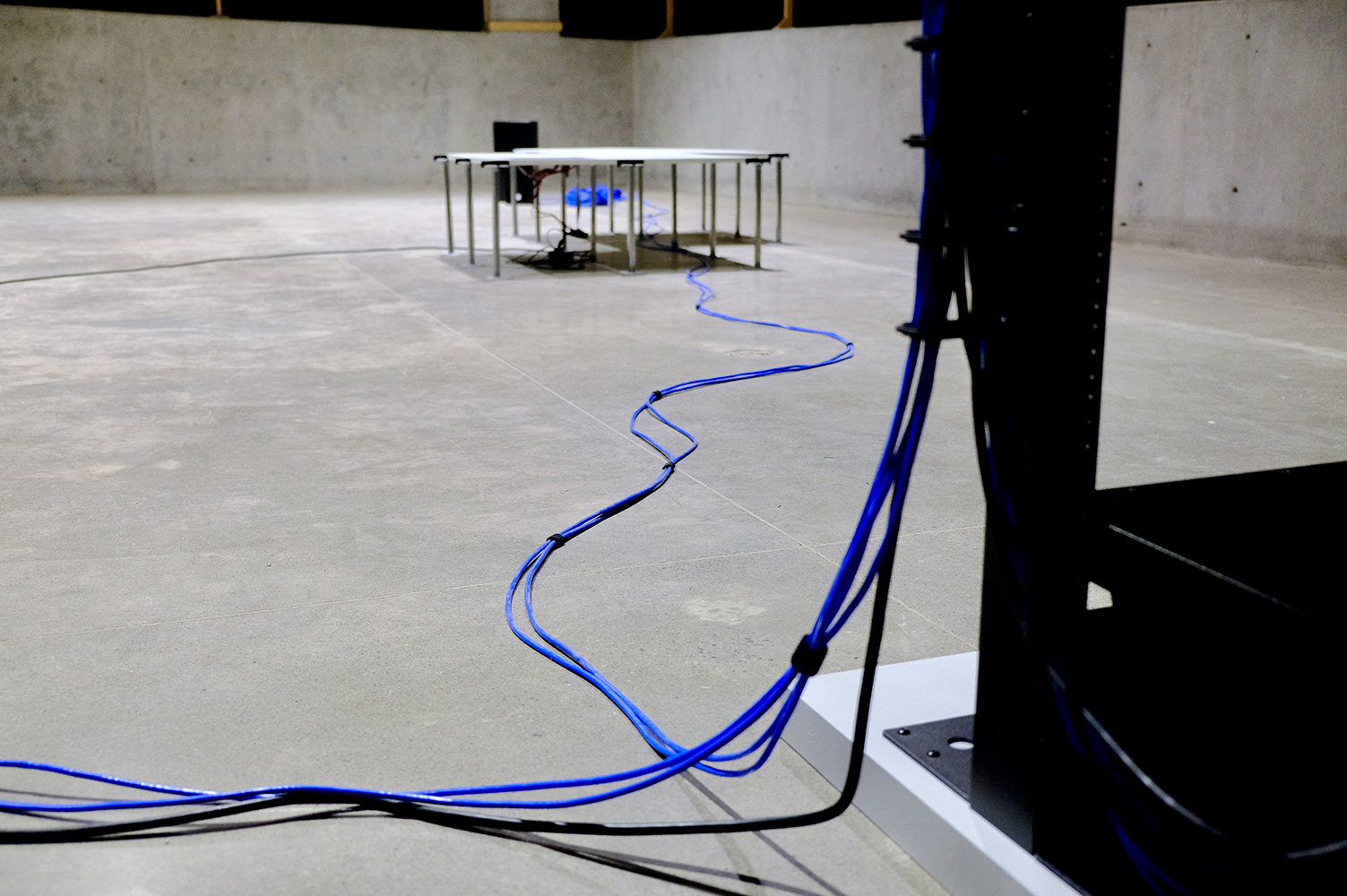 A closeup of the blue ethernet cable exiting the bottom of the server rack and snaking along the floor towards the set of speakers in the distance, running under one of the modular platforms.